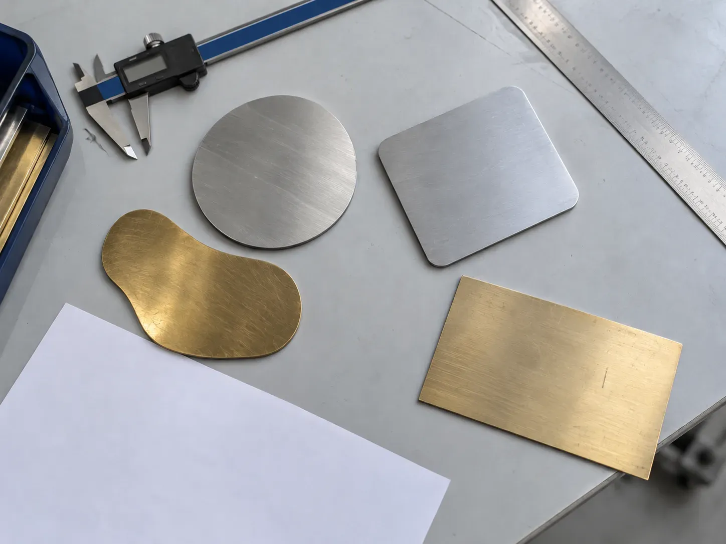 Blank metal shapes with measuring tools on a light grey desk.